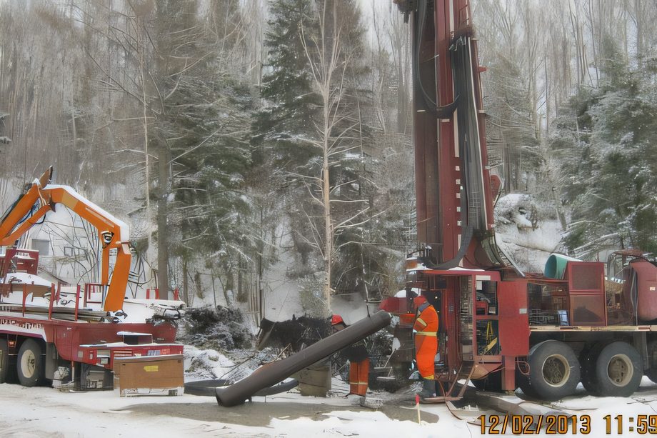 Une foreuse sur camion et une grue sur un chantier de digue enneigé, sapins derrière