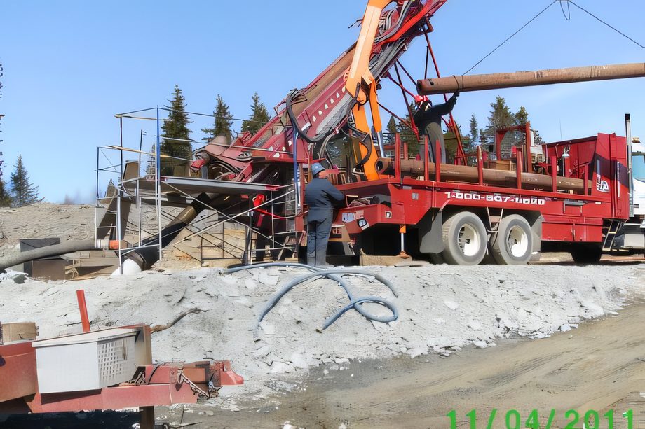Une foreuse rouge avec sa grue sur un chantier de gravier, tubage suspendu